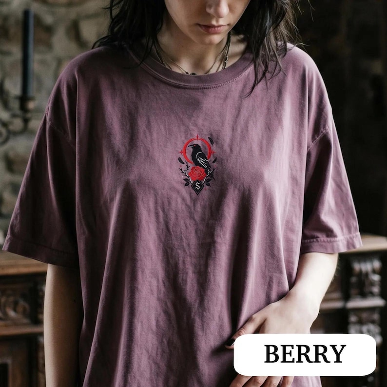 May include: A dusty berry-coloured short-sleeve t-shirt featuring a small, centred graphic on the chest. The design depicts a black crow perched above a red rose, framed by a thin red circle and a small black shield with the letter S inside.