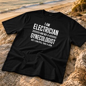 Può includere: Una t-shirt nera a maniche corte con testo bianco che recita I AM ELECTRICIAN AND NOT A GYNECOLOGIST BUT I CAN STILL TAKE A LOOK. La maglia presenta un girocollo ed è realizzata in un tessuto nero a tinta unita con scritte bianche in evidenza.