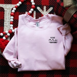 Può includere: Una felpa girocollo rosa chiaro con testo nero sulla parte superiore sinistra del petto che recita FUTURE mrs. *YOUR NAME*. Il capo presenta maniche lunghe e un colletto a coste, mostrando un design semplice con scritte chiare e leggibili.