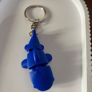 Pode incluir: Um porta-chaves de tubarão articulado azul brilhante com um design de corpo segmentado. O tubarão está preso a um anel de metal prateado por uma corrente curta. A superfície do tubarão mostra linhas de camada de impressão 3D visíveis.