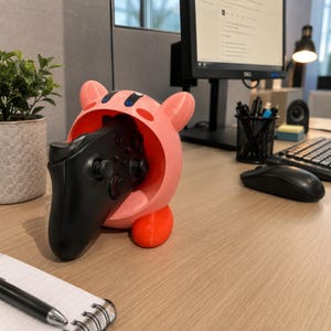 Puede incluir: Un soporte para mando en forma de personaje rosa y redondeado, con dos orejas pequeñas y una boca abierta que sostiene un mando de videojuego negro. El soporte tiene un acabado mate liso con una base de color naranja brillante.