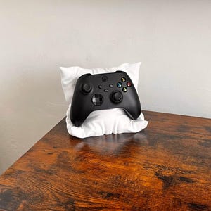 Pode incluir: Um comando de videojogos sem fios preto pousado sobre uma pequena almofada branca, retangular e almofadada. O comando possui dois joysticks pretos, um direcional e quatro botões de ação coloridos marcados com as letras X, Y, B e A.