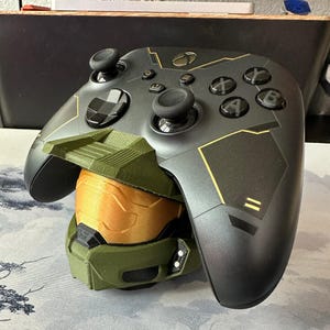 Puede incluir: Un mando de videojuegos inalámbrico negro con detalles dorados y un logotipo de Xbox central, apoyado sobre un soporte impreso en 3D con forma de casco verde y dorado con visera. El mando cuenta con botones, dos joysticks y una cruceta.