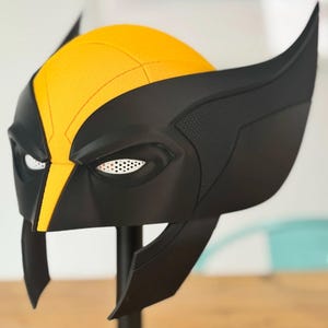 Op de afbeelding: Een geel en zwart superheldenmasker met een centrale gele kam, grote puntige zijvinnen en witte mesh oogbedekkingen. Het masker heeft een gladde matte afwerking met gestructureerde details op de zijpanelen en een gestructureerd, hoekig ontwerp.
