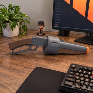 May include: A gray and brown lever-action toy blaster with a rounded barrel housing and an orange tip. The blaster features a brown handle and a gray frame, positioned horizontally on a wooden surface near a computer monitor and a small figurine.