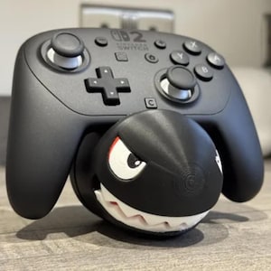 以下が含まれることがあります： 黒いNintendo Switch Proコントローラーが、パックンフラワーのキャラクターを模した白黒の3Dプリント製スタンドの上に置かれています。コントローラーには標準的なボタン、ジョイスティック、中央に白でプリントされたNintendo Switchのロゴがあります。