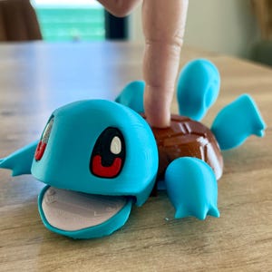 Peut inclure: Une figurine de tortue bleue imprimée en 3D avec une carapace marron, de grands yeux noirs et blancs, et une bouche gris clair. La figurine présente une finition mate lisse sur les membres et la tête bleus, une carapace marron brillante.