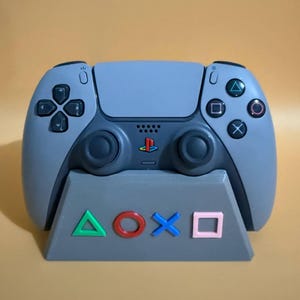 以下が含まれることがあります： 中央にPlayStationロゴ、デュアルアナログスティック、アクションボタンを備えたグレーのワイヤレスビデオゲームコントローラー。コントローラーは、緑の三角形、赤の円、青のX、ピンクの四角形で装飾された、お揃いのグレーの台形スタンドの上に置かれています。