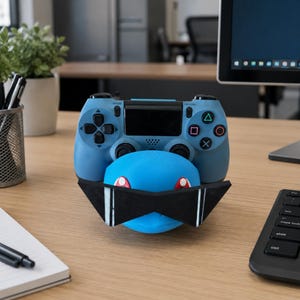 May include: A blue video game controller resting on a blue character-shaped stand that features large, black, angular sunglasses with white accents. The controller has black analog sticks, a black touchpad, and standard gaming buttons.