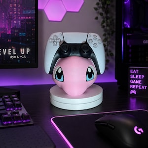 May include: A white and black gaming controller rests on a pink, rounded character-shaped stand with large blue and black eyes. The stand is mounted on a circular white base, positioned on a dark desk surface with a keyboard and mouse nearby.