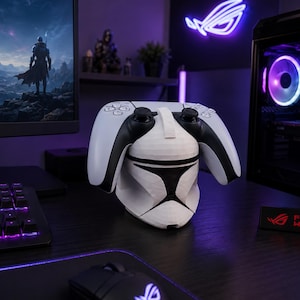 May include: A white and black gaming controller resting on a white, low-poly, geometric clone trooper helmet stand. The stand features a faceted design with black accents around the visor area, providing a stable base for the controller.