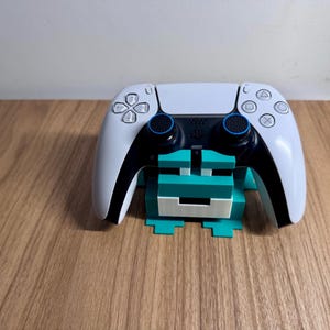 May include: A white and black wireless game controller rests on a teal and white block-style stand. The controller features a central touchpad, two analog sticks with blue rings, and standard action buttons including triangle, circle, cross, and square.