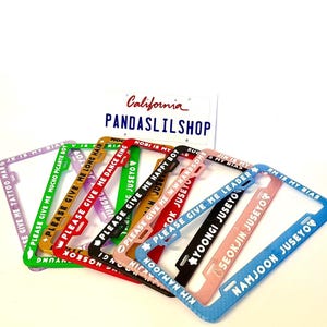 May include: A collection of rectangular license plate frames in various colors including purple, green, yellow, red, black, pink, and blue. Each frame features white text with phrases like PLEASE GIVE ME LONG HAIR and NAMJOON JUSEYO.