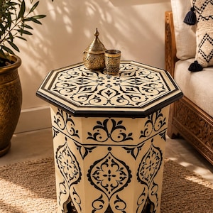 Può includere: Un tavolino ottagonale caratterizzato da una base color crema con intricati motivi floreali e geometrici neri. Il tavolo presenta un piano piatto e aperture ad arco alla base. Un piccolo oggetto decorativo dorato e un bicchiere sono appoggiati sopra.