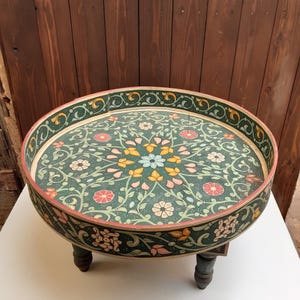 May include: A round wooden table with a low profile and three short, dark-painted legs. The tabletop features a dark green surface decorated with a hand-painted floral pattern in shades of yellow, pink, and light blue, with a matching floral border.