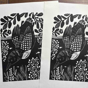 May include: Two identical black and white linocut prints on paper, each featuring a loon swimming in water surrounded by aquatic plants and small fish. The bird is depicted with detailed wing feathers and a patterned neck, set against a dark background.