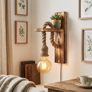 Puede incluir: Un aplique de pared con una placa de madera quemada y un estante horizontal. Una cuerda gruesa de yute natural pasa sobre el estante y sostiene una bombilla de cristal transparente estilo Edison. Una pequeña suculenta en maceta descansa arriba.