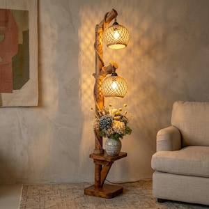 May include: A floor lamp constructed from a vertical wooden post wrapped in thick rope, featuring two spherical woven rattan shades with warm internal lighting. A small wooden shelf holds a ceramic vase filled with a bouquet of dried flowers.