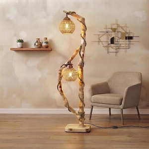 Peut inclure: Un lampadaire composé d'une structure en branche de bois clair naturel avec deux abat-jours sphériques en rotin tressé. La branche est entourée d'une corde épaisse et la lampe repose sur un socle rectangulaire en bois avec un cordon noir.