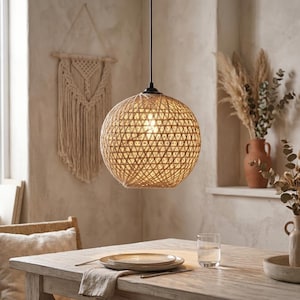Puede incluir: Una lámpara colgante esférica con un exterior de yute o cuerda trenzada de color tostado con un patrón geométrico triangular. La luz está suspendida por un cable negro fino y emite un brillo cálido desde el centro, resaltando su textura.