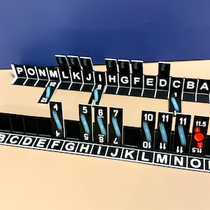 以下が含まれることがあります： 白い文字と数字が記された2つの黒いプラスチック製ゲームラック。手前のラックにはAからPまでの文字が表示され、4、5、6、7、10、11、11、11.5の数字が書かれた垂直のタイルが並んでいます。奥のラックにはAからPまでの文字が表示されています。