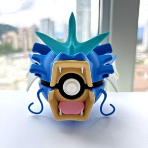 May include: A 3D printed figurine of a blue sea creature with a large tan jaw, white fangs, and a pink tongue. The creature features a teal crest on its head, white side fins, and two curved blue whiskers. A black and white sphere is inside the mouth.