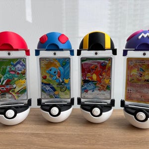 May include: Four vertical clear plastic trading card display cases, each mounted on a white and black spherical base. The top caps are coloured red, blue with red spots, black with yellow stripes, and purple with a white M logo, holding Pokémon cards.