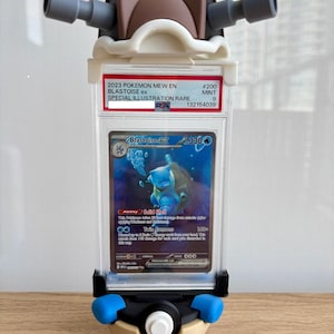 May include: A PSA graded 2023 Pokemon Mew EN Blastoise ex #200 Mint 9 trading card encased in a clear plastic slab. The slab is mounted in a custom 3D-printed display stand shaped like a Blastoise shell with two grey cannons and a Pokeball base.