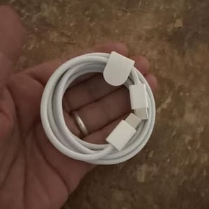 May include: A white charging cable with USB-C connectors, coiled neatly. The cable is held in a hand, with the connectors and cable ends visible. The background is a neutral, textured surface.