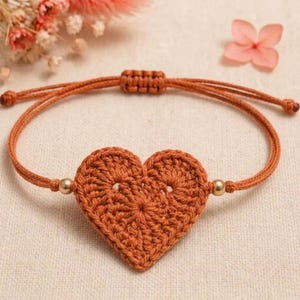 May include: A rust-colored crochet heart bracelet featuring a central heart-shaped motif made of yarn, two small metallic gold-toned beads on either side, and an adjustable sliding knot closure made from the same rust-colored cord material.