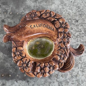 May include: A brown ceramic dish shaped like a fish, featuring a relief of flowers surrounding a central green glazed basin. The word CALIFORNIA is embossed in black lettering on the upper body of the fish, which has a textured finish.