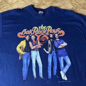 Könnte beinhalten: Ein marineblaues Kurzarm-T-Shirt mit einem Grafikdruck der Musikgruppe The Oak Ridge Boys. Das Design zeigt vier Personen, die in einer Reihe stehen und Freizeitkleidung tragen, mit dem Bandnamen in gelbem und orangefarbenem Text darüber.