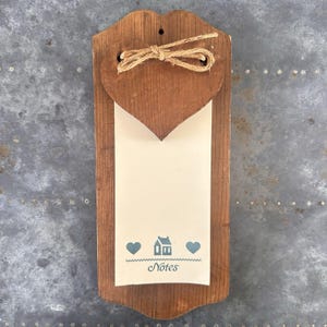 May include: A rectangular wooden wall-mounted note holder with a dark brown finish. A wooden heart cutout is attached at the top with a jute twine bow. A cream-colored paper pad features a small house icon, two hearts, and the word Notes.
