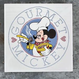 May include: A square white ceramic tile featuring a circular graphic of Mickey Mouse dressed as a chef, wearing a white hat and yellow apron, holding a bowl of pasta. The text GOURMET MICKEY is written in a cross-stitch style around the circle.