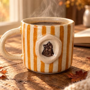 Puede incluir: Una taza de cerámica con rayas verticales de color naranja y crema, que presenta un medallón circular en relieve en la parte frontal con una ilustración detallada en marrón de una cara de gato. La taza contiene un líquido oscuro y desprende vapor.