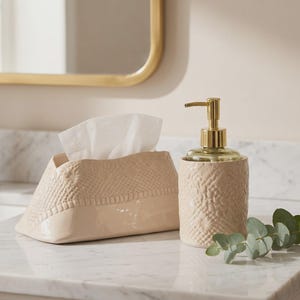 Op de afbeelding: Een beige keramieken tissueboxhouder en een bijpassende zeepdispenser met een goudkleurige pomp. Beide items hebben een gestructureerd, reliëfpatroon aan de bovenzijde en een gladde, glanzende afwerking aan de onderzijde met witte tissues.