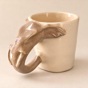 May include: A ceramic mug with a glossy cream-colored finish and a sculpted handle shaped like an elephant head. The elephant handle is finished in a smooth, light brown glaze, featuring detailed ears, a trunk, and tusks that form the grip.
