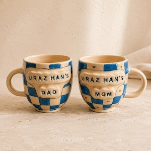 Puede incluir: Dos tazas de cerámica con un patrón de cuadros azul y crema. Cada taza presenta un panel central en relieve con el texto URAZ HAN'S DAD en una y URAZ HAN'S MOM en la otra. Las tazas tienen un acabado brillante y pequeños detalles de corazones dorados.