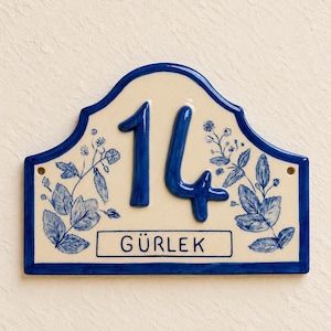 Pode incluir: Uma placa de casa retangular em cerâmica com topo curvo, fundo creme e borda azul-escura. A placa exibe o número 14 em grandes dígitos azuis, o texto GÜRLEK dentro de uma caixa azul e ilustrações florais na cor azul.