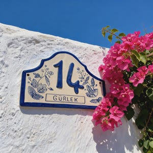 May include: A rectangular ceramic house number plaque with a curved top edge, featuring the number 14 and the text GÜRLEK in blue. The plaque has a cream-colored background with blue floral illustrations and a blue border, mounted on a white wall.