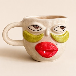 May include: A ceramic mug with a beige, uneven surface shaped like a human face. It features two dark brown eyes, two bright green under-eye patches, and prominent, glossy red lips. The mug has a single curved handle on the left side.