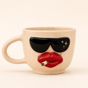 Pode incluir: Uma caneca de cerâmica bege com um design de rosto em relevo tridimensional. A caneca apresenta óculos de sol pretos brilhantes com detalhes de estrelas brancas, lábios vermelhos brilhantes e um pequeno cigarro saliente entre os lábios.