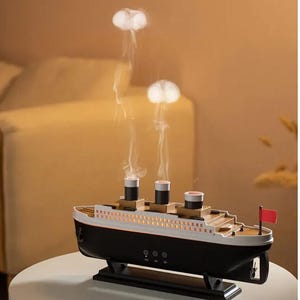 Puede incluir: Un humidificador con forma de barco en blanco y negro con tres chimeneas que emiten anillos de vapor blancos. El navío cuenta con una bandera roja en la popa, casco negro, cubierta blanca y tres botones de control en el lateral sobre una base.