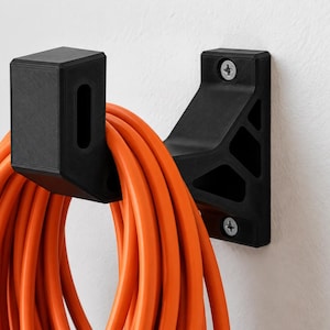 Op de afbeelding: Een zwarte wandhaak met een geometrische, open steunstructuur, bevestigd met twee zilverkleurige schroeven. Een lang, feloranje verlengsnoer is opgerold en hangt netjes over de haak voor een georganiseerde opslag van kabels.