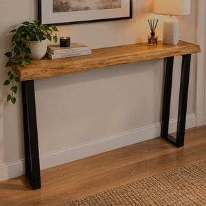 Pode incluir: Uma mesa consola comprida com um tampo de madeira castanho-claro de bordo natural e duas pernas de metal preto em forma de U. A superfície da mesa contém uma planta verde em vaso, dois livros empilhados, uma vela preta e um difusor.