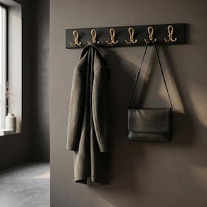 Könnte beinhalten: Eine rechteckige, schwarze Wandgarderobe mit sechs goldfarbenen Metallhaken in geschwungenem Design. Ein dunkelgrauer Wollmantel hängt an einem Haken und eine schwarze Umhängetasche aus Leder hängt an einem weiteren Haken.