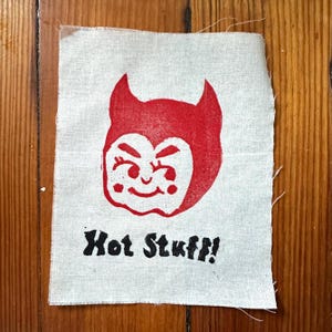 May include: A rectangular piece of off-white fabric featuring a red screen-printed graphic of a cartoon devil face with horns and a smiling expression. Below the devil face, the text "Hot Stuff!" is printed in bold, black, capital letters.