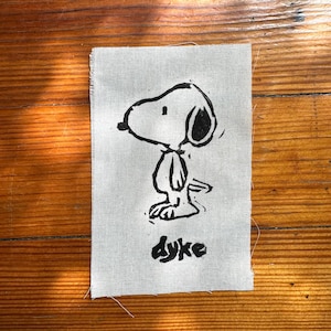 May include: A rectangular piece of light gray fabric featuring a black ink block print of a cartoon dog in profile. The dog has a large head, floppy ear, and simple body lines. Below the dog, the word dyke is printed in a bold, hand-drawn style.