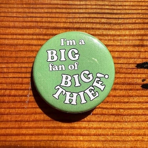 May include: A round pinback button with a light green background. The text printed in white with a black outline reads: I'm a BIG fan of BIG THIEF! The button has a smooth, glossy finish and a circular shape, featuring bold, playful typography.