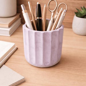 Peut inclure: Un pot à crayons cylindrique de couleur lavande avec des cannelures verticales, contenant divers stylos, crayons et une paire de ciseaux en métal doré. Le récipient présente une finition mate et est utilisé pour ranger des fournitures.